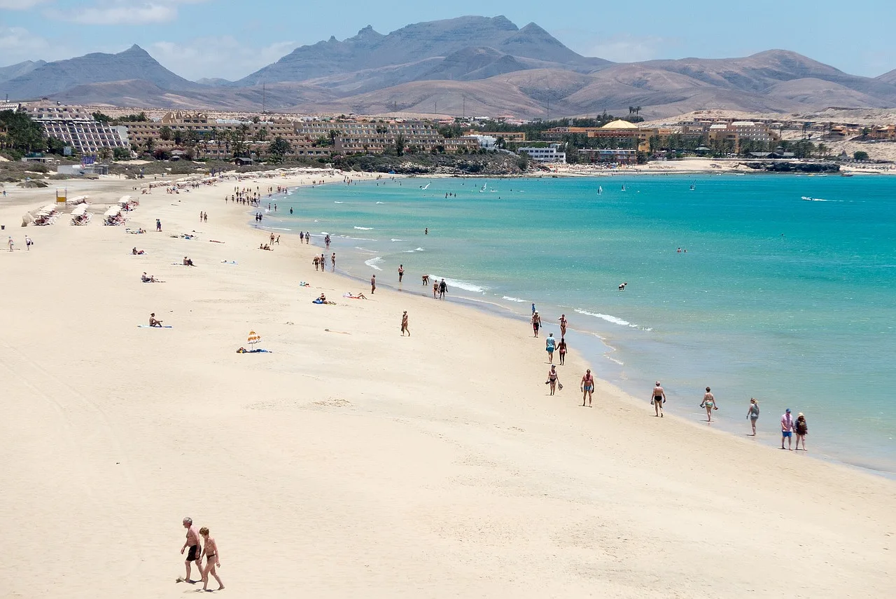 piaszczysta plaża, turyści oraz resorty na wybrzeżu Fuerteventura, góry w tle