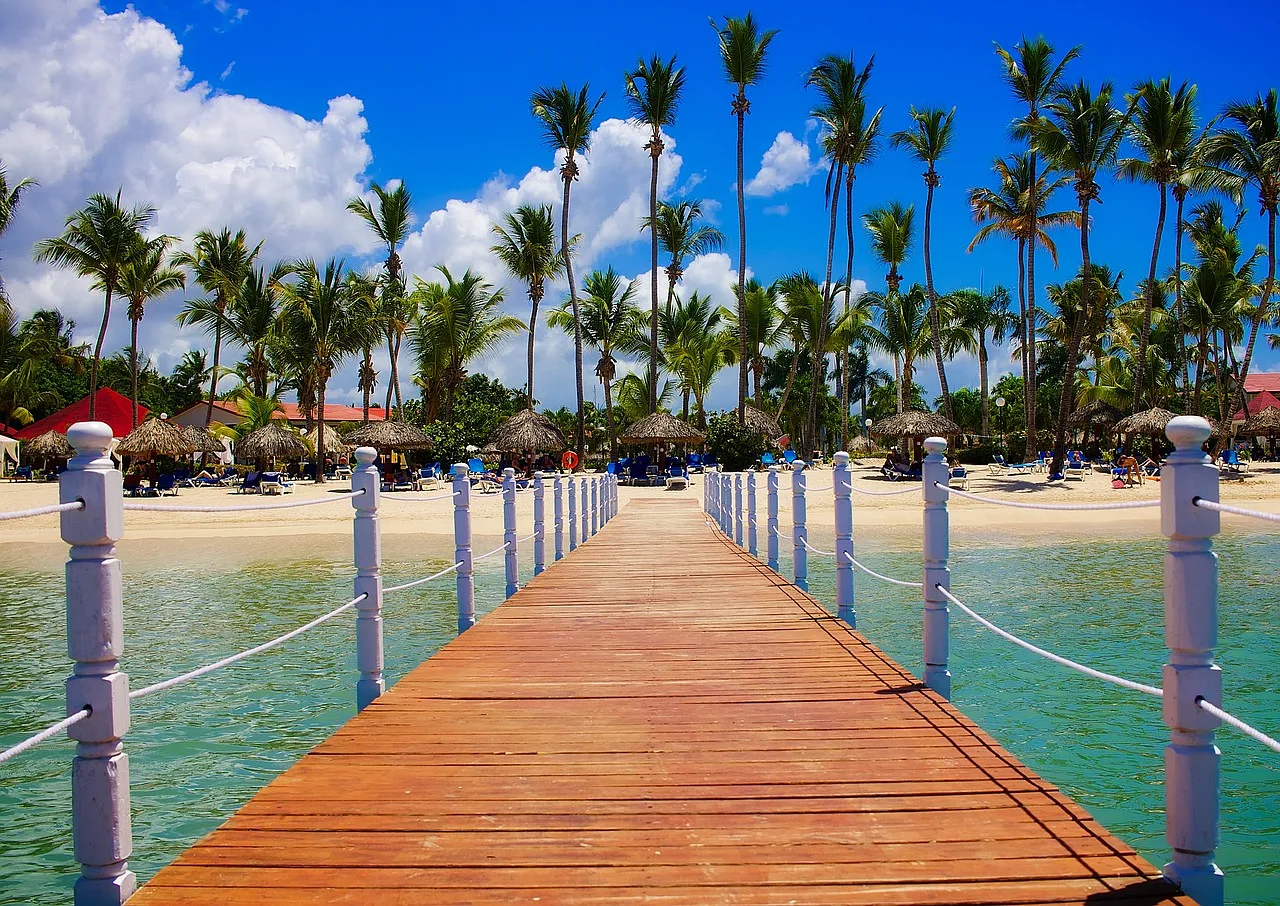pomarańczowy pomost z białymi poręczami oraz piaszczysta plaża z zielonymi palmami w tle