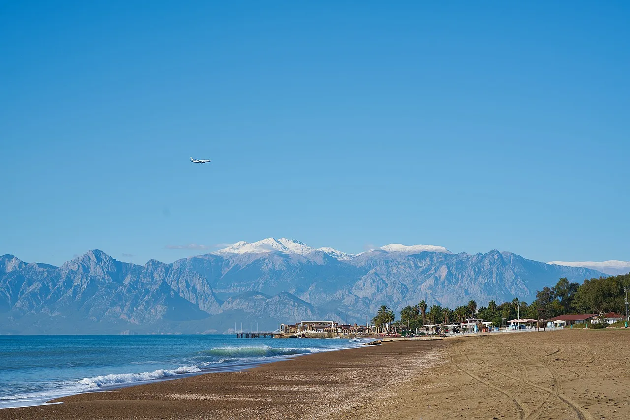 Widok na plażę w Antalya, Turcja, z górami Taurus w tle i samolotem lecącym nad wybrzeżem.