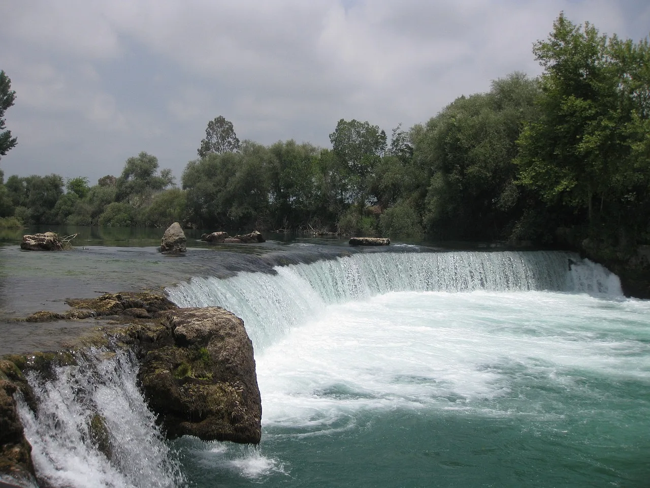 Zdjęcie Wodospadu Manavgat w Turcji, przedstawiające szeroką kaskadę wody spływającą po płaskim kamiennym progu, otoczoną zielonymi drzewami i bujną roślinnością.