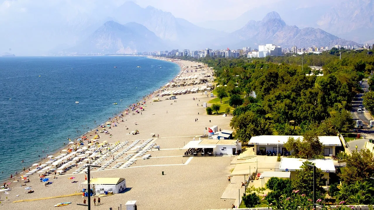 Widok na plażę w Antalyi, Turcja, z widocznymi górami w tle, leżakami i parasolami na plaży oraz turystami korzystającymi z letniego wypoczynku.