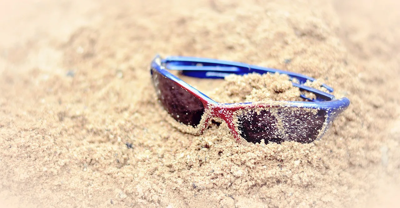 Okulary przeciwsłoneczne leżące na piasku, pokryte drobinkami, symbolizujące letni wypoczynek na plaży.
